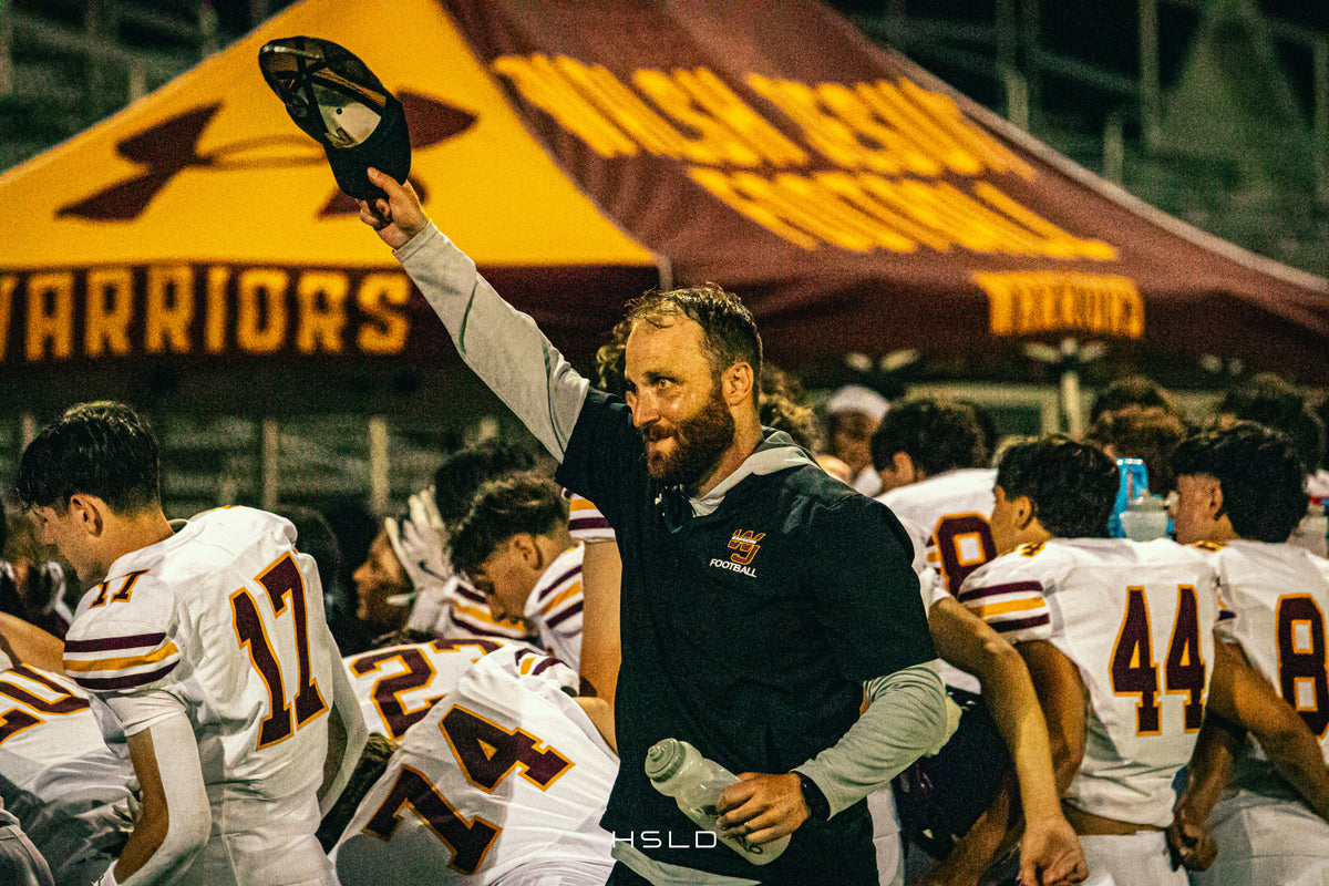 Football coach raising his cap beside players on the sideline.